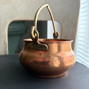 Può includere: Un piccolo vaso color rame con un manico in ottone. Il vaso ha un corpo arrotondato e un bordo leggermente svasato. Il manico è arcuato e attaccato al vaso con staffe in ottone. Il vaso è appoggiato su una superficie scura.