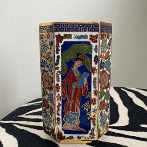 Könnte beinhalten: Eine achteckige Cloisonné-Vase mit detailliertem Design. Die Vase zeigt ein zentrales Feld mit einer Frau in rotem Gewand, umgeben von floralen und geometrischen Mustern in Blau, Weiß und Gold. Die Vase steht auf einer zebraartig gestreiften Oberfläche.