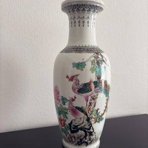 Op de afbeelding: Witte keramische vaas met een gedetailleerd vogel- en bloemdesign. De vaas heeft een lange hals met een decoratieve rand en een uitlopende basis. De vogel heeft roze, teal en groene veren. Een decoratief woonaccessoire.