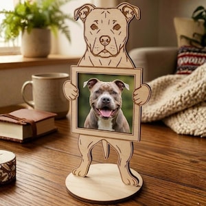 Pode incluir: Uma moldura de madeira em forma de cão, segurando uma foto de um cão sorrindo. A moldura é feita de madeira clara e tem uma base circular. As características do cão são gravadas na madeira.