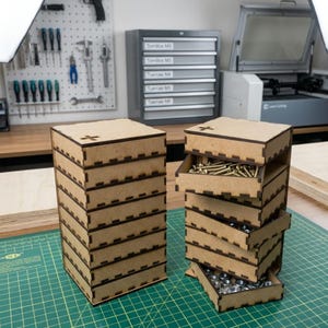 May include: Two light brown wooden storage towers with multiple stacked drawers featuring interlocking finger joints. One tower is closed, while the other has three drawers partially extended, revealing metal screws and nuts inside the compartments.