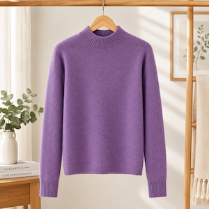 May include: A solid purple long-sleeve sweater featuring a ribbed mock neck collar, ribbed cuffs, and a ribbed hem. The garment is constructed with a soft-textured knit fabric and is displayed hanging on a wooden hanger against a neutral background.