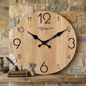 May include: A round wall clock with a light wood grain finish, featuring large black Arabic numerals and black metal hands. The clock face displays the text "est 1765 vintage clocks" and "QUARTZ CLOCK" in small black lettering below the number 12.
