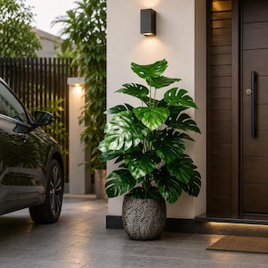 May include: A large, vibrant green Monstera plant with broad, split leaves housed in a dark, textured, cylindrical planter with a repeating leaf-like pattern, positioned on a tiled floor near a modern entrance with a dark wooden door.