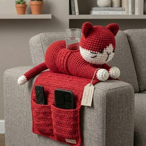 Puede incluir: Un organizador de brazo de sofá con forma de gato rojo tejido a ganchillo, con dos bolsillos para mandos a distancia y teléfonos. El gato tiene detalles blancos en la cara y las patas, y una cola con punta blanca. Un vaso de agua está en la espalda del gato.
