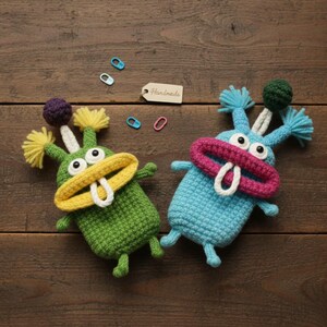 May include: Two handmade crochet monster toys in green and blue, with large eyes, open mouths, and pom-pom accents. The green monster has a yellow mouth and the blue monster has a pink mouth. A tag that says "Handmade" is visible.