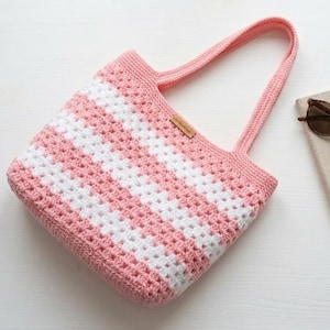 May include: A pink and white striped crochet tote bag with two handles. The bag has a rectangular shape and a small brown tag. Sunglasses and a book are next to the bag on a white surface.