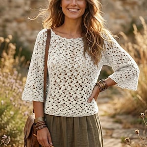 May include: A cream-colored, open-weave crochet top with three-quarter sleeves. The top has a round neckline and a loose, airy design. The model is wearing a brown leather shoulder bag and a green skirt.