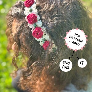 Può includere: Una fascia per capelli all'uncinetto fatta a mano con rose rosse vibranti e foglie verdi. La corona floreale è posta su capelli ricci scuri. L'immagine include il testo "PDF PATTERN + VIDEO" e "ENG (US)" e "IT".