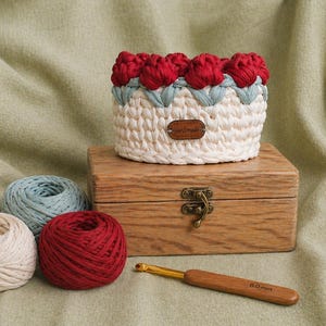 May include: A handmade crochet basket with red flower accents, sitting on a wooden box with a clasp. Three yarn balls in cream, blue, and red are nearby, along with a crochet hook. The basket is white with red and blue details.