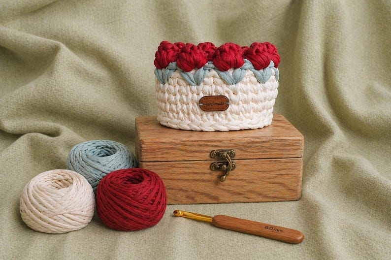 May include: A handmade crochet basket with red flower accents, sitting on a wooden box with a clasp. Three yarn balls in cream, blue, and red are nearby, along with a crochet hook. The basket is white with red and blue details.