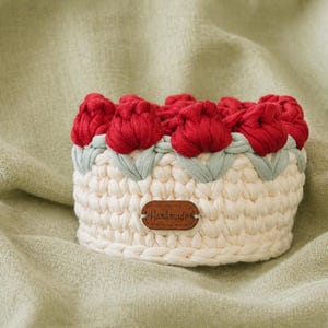 May include: A handmade, cream-colored crochet basket with red tulip-shaped accents. The basket features a light green trim and a small, brown tag that reads "Handmade". The basket is sitting on a light green fabric.