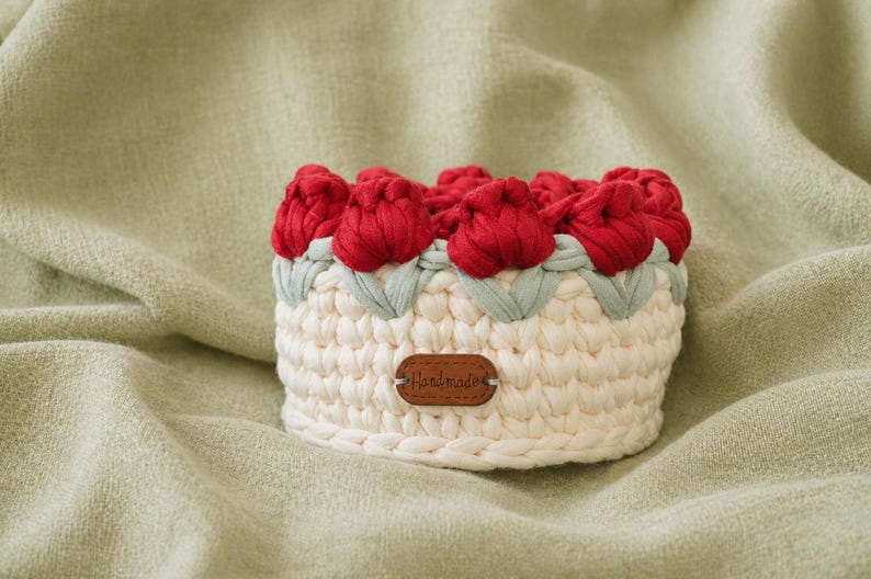 May include: A handmade, cream-colored crochet basket with red tulip-shaped accents. The basket features a light green trim and a small, brown tag that reads "Handmade". The basket is sitting on a light green fabric.