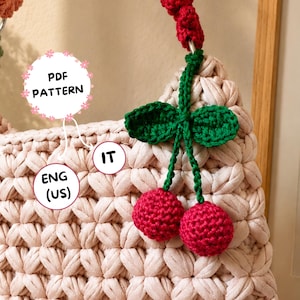 Può includere: Una borsa all'uncinetto rosa chiaro con un design a ciliegia. La borsa presenta un motivo a punto strutturato e un manico decorato con ciliegie all'uncinetto rosse e foglie verdi. Il testo "PDF PATTERN" e "ENG (US)" sono visibili.