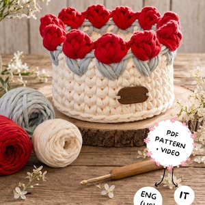 May include: A handmade crochet basket with red flower accents. The basket is cream-colored with a gray trim and red flowers around the top. A wooden crochet hook and yarn balls in red, cream, and gray are also visible.