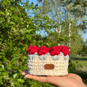 May include: A handmade, cream-colored crochet basket with red flower accents and a gray trim. The basket has a small, brown leather tag that reads "Handmade". The basket is held in a person's hand.