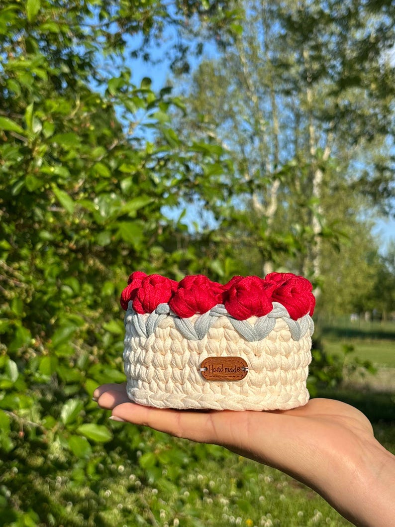 May include: A handmade, cream-colored crochet basket with red flower accents and a gray trim. The basket has a small, brown leather tag that reads "Handmade". The basket is held in a person's hand.