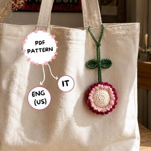 Può includere: Una borsa tote color crema con un ciondolo a forma di fiore all'uncinetto. Il fiore ha un centro bianco, uno strato intermedio rosa e uno strato esterno rosso scuro. Il ciondolo è attaccato al manico della borsa con uno stelo verde all'uncinetto. Il testo sull'immagine dice "PDF PATTERN" e "ENG (US)".
