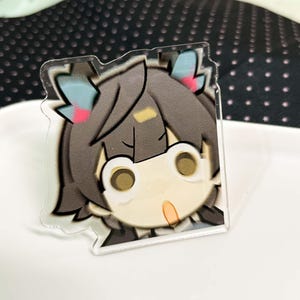 May include: A clear acrylic charm featuring a stylized cartoon character with dark brown hair, large circular eyes, and small blue and pink accents in the hair. The character has a surprised expression with an open mouth and a small yellow mark on the forehead.