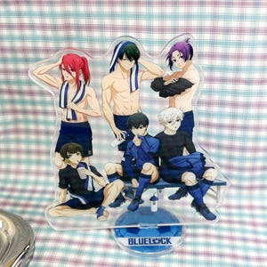 May include: A clear acrylic standee featuring five anime characters from Blue Lock. The characters are shirtless or wearing athletic gear, with some holding towels. The base is circular and blue with the text BLUELOCK printed in white.