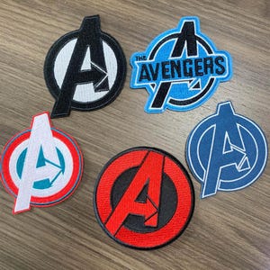 Op de afbeelding: Vijf geborduurde patches met het Avengers-logo in verschillende kleuren. De patches bevatten de letter "A" in zwart, rood, wit en blauw, met de woorden "The Avengers" op één patch. Deze patches zijn geschikt voor kleding of accessoires.