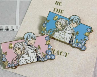 Könnte beinhalten: Zwei rechteckige Emaille-Anstecknadeln mit Anime-Figuren mit hellem Haar und weißen Umhängen. Eine Anstecknadel hat einen hellrosa Hintergrund, die andere einen hellblauen. Beide sind mit kleinen blauen Blumen und gelben Schmetterlingen verziert.