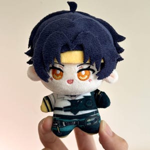 Peut inclure: Une petite poupée en peluche avec des cheveux bleu foncé, un bandeau jaune et des yeux brodés orange. La poupée porte une chemise blanche avec des accents noirs, un gilet sombre et un pantalon bleu sarcelle avec des détails imprimés.