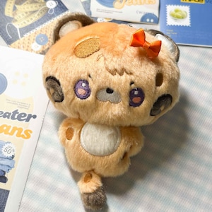 May include: A plush toy shaped like a small animal with light orange fur, dark brown spots, and a white belly patch. It features large purple embroidered eyes, a small gray nose, and a bright orange satin bow attached to the top of its head.
