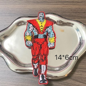 Op de afbeelding: Geborduurde patch met een gespierde superheld in een loophouding. De patch is voornamelijk rood, met gele en blauwe accenten. De patch meet 14 cm bij 6 cm.