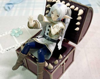 May include: A figurine of a white-haired elf character with pointed ears, wearing a white coat with gold trim and dark blue pants, sitting inside a brown treasure chest with sharp, cream-colored teeth lining the interior edges of the lid and base.