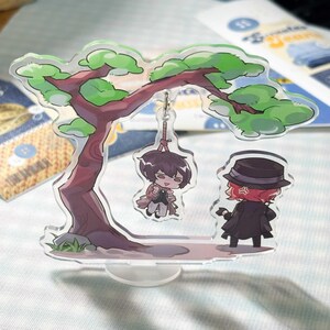 May include: An acrylic standee featuring a stylised tree with green foliage and a brown trunk. A small character figure hangs from a branch by a rope, while a second character wearing a black hat and coat stands on the ground below the tree.