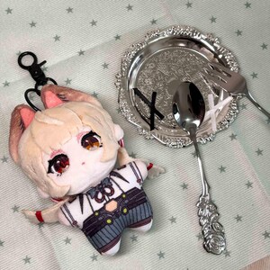 May include: A plush doll with blonde hair, cat ears, and red eyes, wearing a white and grey outfit with a black keychain attachment. Next to it is a silver-toned decorative plate with an intricate pattern, a silver spoon, and a silver fork.
