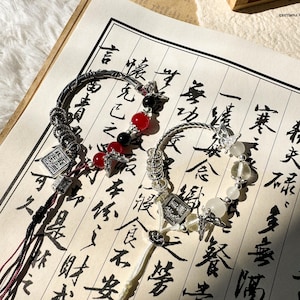 Puede incluir: Dos pulseras ornamentadas con elementos metálicos plateados y cuentas de colores, incluyendo rojas, negras y blancas. Cada pulsera presenta un dije decorativo y una borla, sobre una hoja de papel con caligrafía.