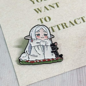 May include: An enamel pin featuring a white-haired elf character with pointed ears, wrapped in a white cloak, sitting on a red and white checkered blanket. A small white creature and a black cat are beside the character on a patch of green grass.