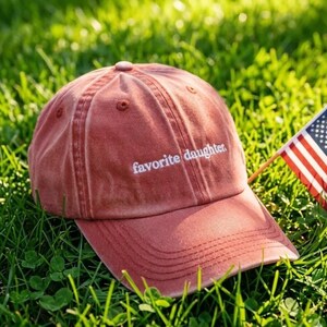 May include: A dusty rose baseball cap with the embroidered text "favorite daughter." The cap is resting on a bed of green grass, with a small American flag in the background.