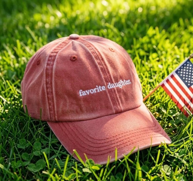 May include: A dusty rose baseball cap with the embroidered text "favorite daughter." The cap is resting on a bed of green grass, with a small American flag in the background.
