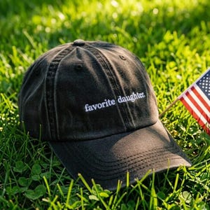 May include: A dark gray baseball cap with the embroidered text "favorite daughter" in white. The cap is resting on green grass next to a small American flag.