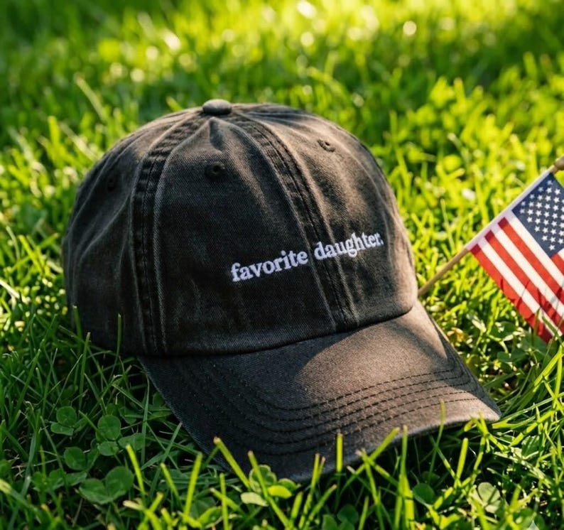 May include: A dark gray baseball cap with the embroidered text "favorite daughter" in white. The cap is resting on green grass next to a small American flag.