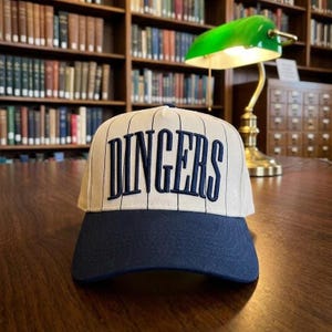 May include: A baseball cap featuring a cream-colored crown with thin vertical black pinstripes and the word DINGERS embroidered in bold navy blue lettering across the front. The cap has a solid navy blue brim and a structured five-panel design.