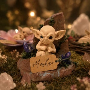 Può includere: Statuina beige di una creatura con grandi orecchie e un'espressione intensa, appoggiata su una formazione rocciosa. Un'etichetta con il nome "Morkai" è attaccata. La scena include altre statuine ed elementi decorativi.