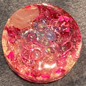 May include: A round, clear resin pendant with pink and iridescent glitter and bubbles. The pendant has a pink and white swirl pattern.