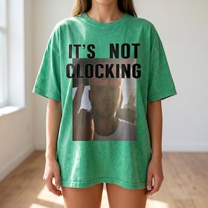 May include: A green t-shirt with the text "IT'S NOT CLOCKING" in black block letters. The shirt features a faded image of a person wearing a pink hat. The shirt is a casual, oversized style.