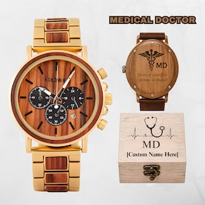 May include: A gold and wood watch with a chronograph dial, a second watch with a brown leather strap and a caduceus engraving, and a wooden box with a stethoscope graphic and the text MD and Custom Name Here, labeled MEDICAL DOCTOR.