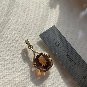 May include: A gold pendant featuring an oval-cut, amber-colored gemstone set in a decorative frame with a bail at the top. A metal ruler is positioned next to the pendant, showing the gemstone measures approximately 1.5 centimeters in length.