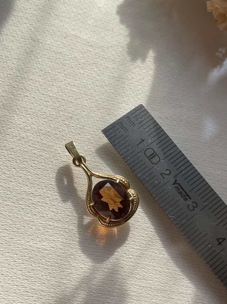 May include: A gold pendant featuring an oval-cut, amber-colored gemstone set in a decorative frame with a bail at the top. A metal ruler is positioned next to the pendant, showing the gemstone measures approximately 1.5 centimeters in length.
