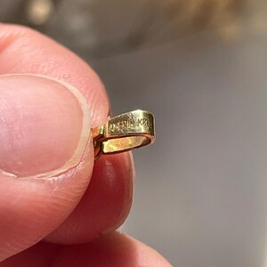 May include: A small, gold-colored metal jewelry bail with a polished finish. The surface of the bail features a stamped inscription that reads AMERIK 22K, indicating the metal purity and origin. The piece has a curved, rectangular loop shape.