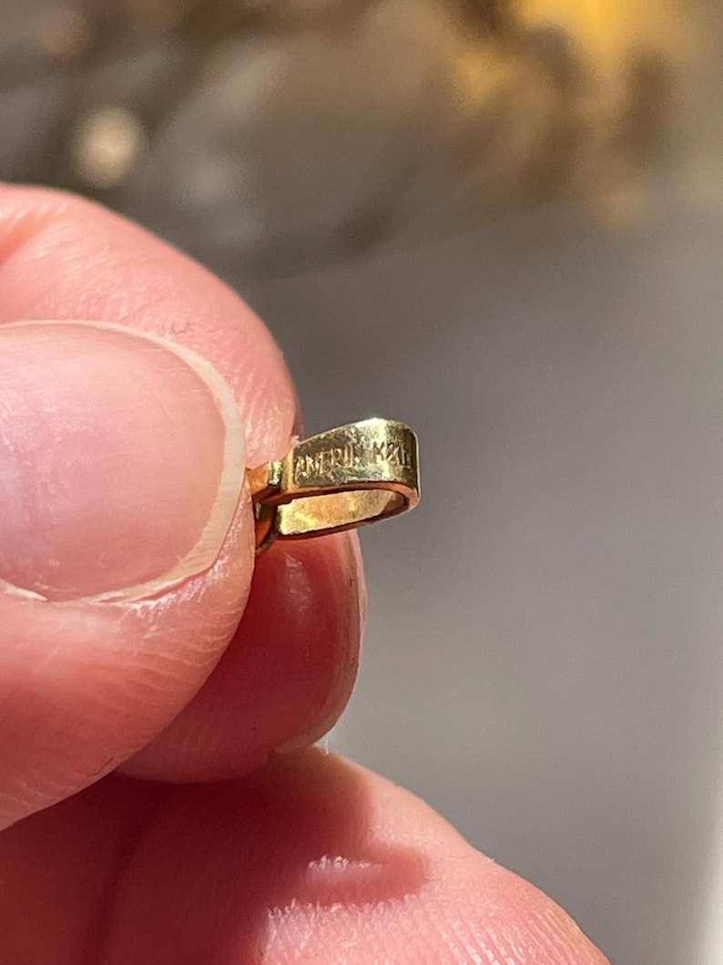 May include: A small, gold-colored metal jewelry bail with a polished finish. The surface of the bail features a stamped inscription that reads AMERIK 22K, indicating the metal purity and origin. The piece has a curved, rectangular loop shape.