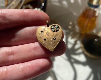 May include: A heart-shaped gold-tone pendant featuring an engraved floral pattern with three small dark inset stones and a single light blue enamel flower accent in the upper right corner, complete with a small metal loop at the top for attachment.