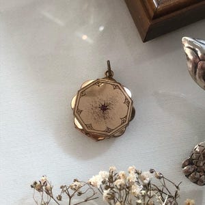 May include: A gold-toned metal locket with an octagonal front panel featuring an engraved border and a small, centered, star-set red gemstone. The locket has a scalloped edge design and a small metal loop at the top for attachment to a chain.