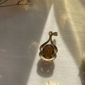 May include: A gold-toned pendant featuring an oval-shaped, translucent amber-colored gemstone set within a curved, open-work metal frame. The pendant includes a small circular bail at the top for attachment to a chain or cord.